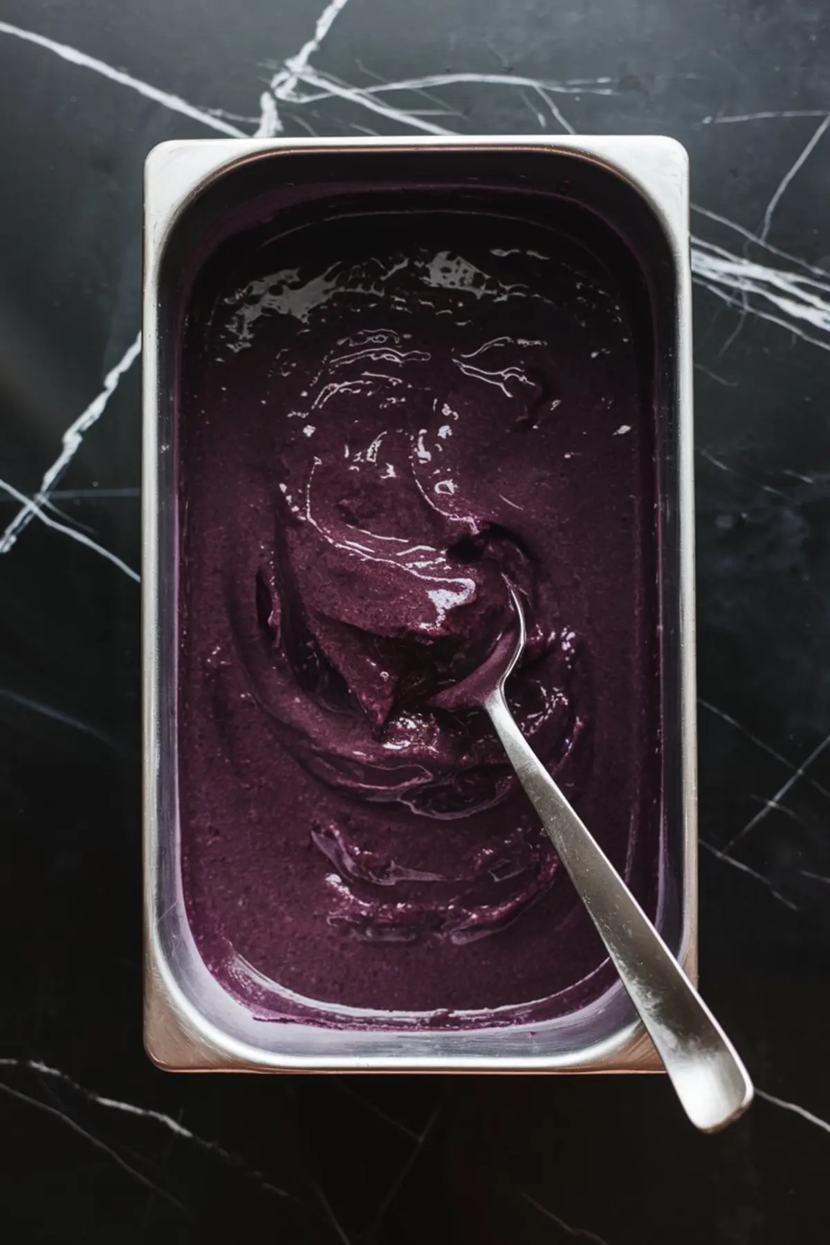 Overhead view of a metal container filled with freshly blended berry lemonade sorbet, with a spoon partially submerged, highlighting the smooth, thick consistency and rich berry hue.