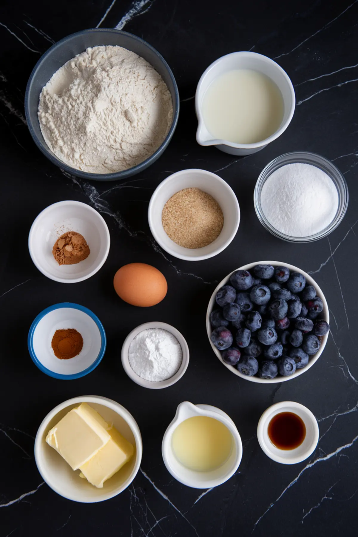 A flat lay of ingredients for blueberry cinnamon rolls on a black marble countertop. Ingredients include a bowl of flour, sugar, brown sugar, cinnamon, an egg, butter, vanilla extract, milk, baking powder, and a bowl of fresh blueberries.