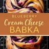 Blueberry cream cheese babka with a golden brown crust, swirls of blueberry filling, and a dusting of powdered sugar. The twisted dough design reveals layers of soft bread and rich filling. The babka sits on a cooling rack over a black marble surface, with a brown text overlay that reads “Blueberry Cream Cheese Babka.”