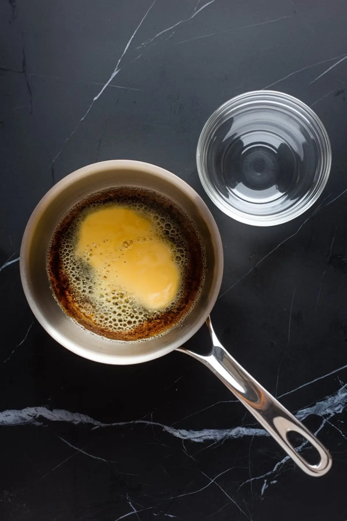 Saucepan with browned butter bubbling on a dark marble surface, next to an empty glass bowl for mixing.