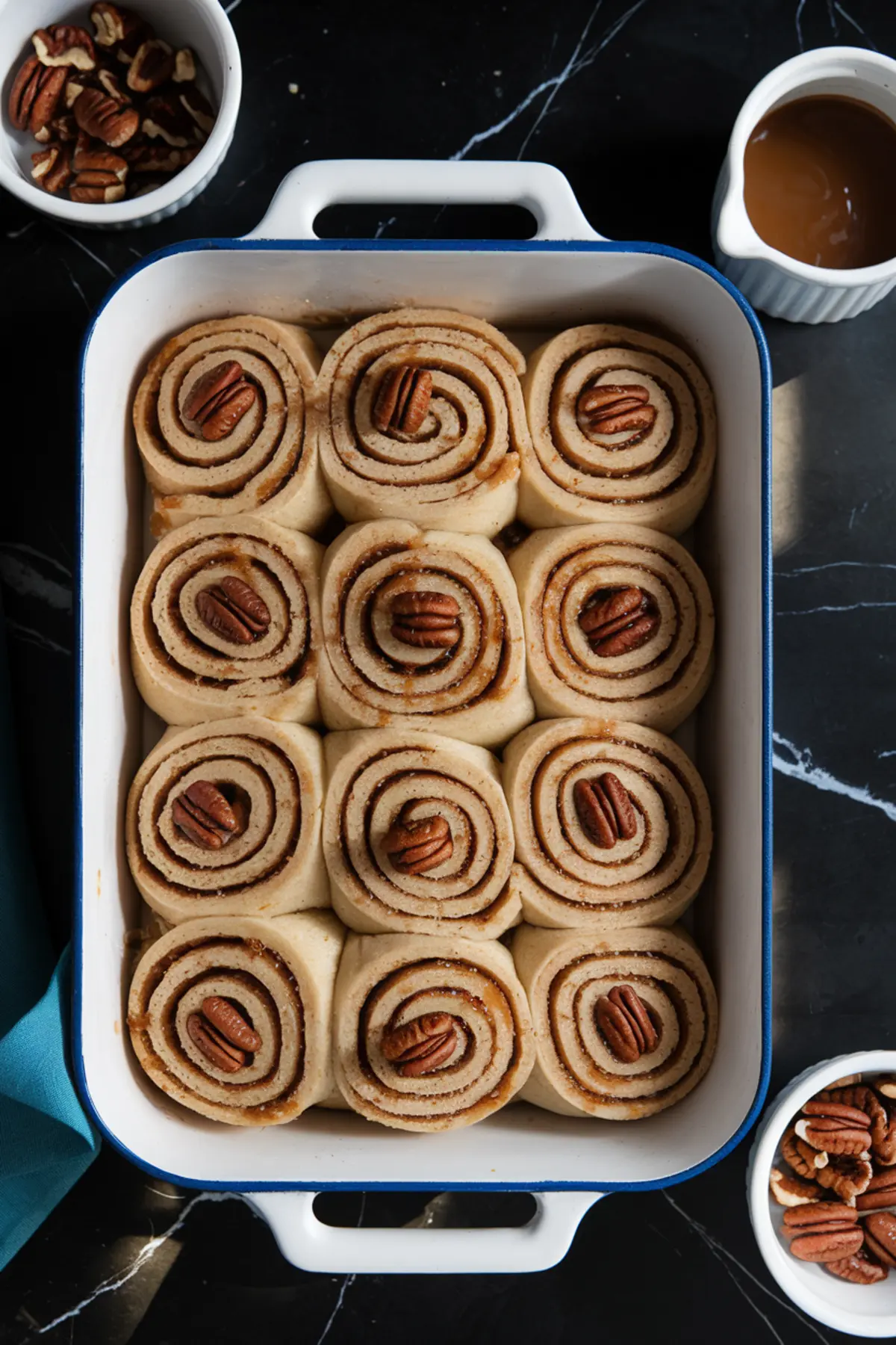 A baking dish filled with unbaked spiral sticky buns arranged in rows, each topped with a pecan half, with bowls of pecans and caramel sauce in the background on a dark countertop.