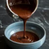 Close-up of warm chocolate ganache being poured from a saucepan into a textured blue bowl. The ganache forms ripples as it collects, creating a smooth, shiny swirl on the surface.