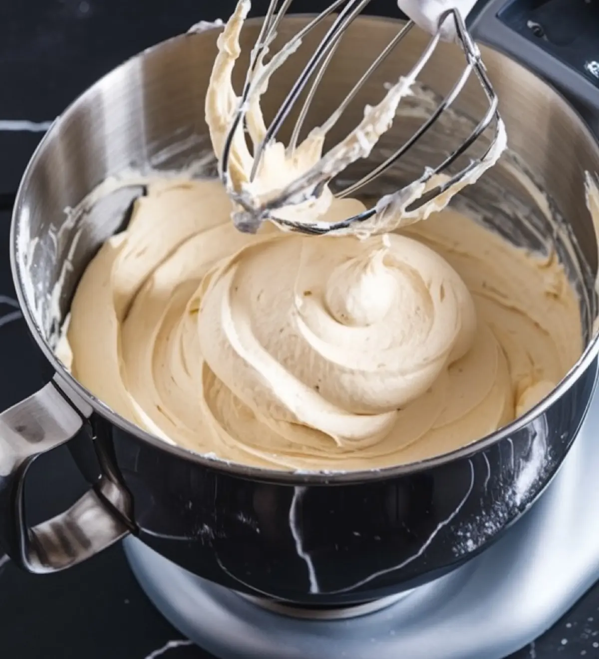 A mixing bowl with smooth, freshly whipped buttercream frosting. A stand mixer whisk attachment is coated with the creamy frosting, set against a black marble countertop.