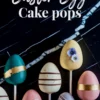 Easter egg cake pops display egg-shaped treats on sticks set against a black marble background. Covered in pastel shades of teal, pink, and beige, each cake pop is adorned with gold foil bands and small edible flowers.