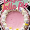 A pastel pink Easter Jello pie with a graham cracker crust in a white fluted pie dish. The pie is decorated with whipped cream swirls, edible flowers in shades of purple and yellow, and tiny sprinkles. Bold pink and teal text overlays the image, reading “Easter Jello Pie.” The background includes pastel-colored napkins and a dark marble surface.