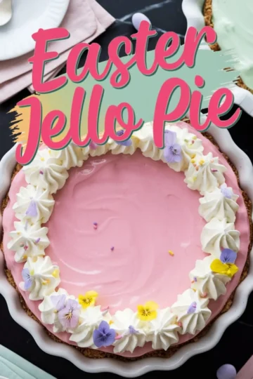 A pastel pink Easter Jello pie with a graham cracker crust in a white fluted pie dish. The pie is decorated with whipped cream swirls, edible flowers in shades of purple and yellow, and tiny sprinkles. Bold pink and teal text overlays the image, reading “Easter Jello Pie.” The background includes pastel-colored napkins and a dark marble surface.