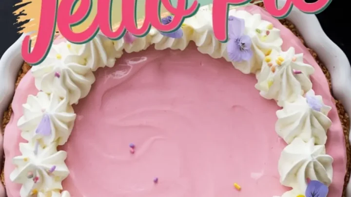 A pastel pink Easter Jello pie with a graham cracker crust in a white fluted pie dish. The pie is decorated with whipped cream swirls, edible flowers in shades of purple and yellow, and tiny sprinkles. Bold pink and teal text overlays the image, reading “Easter Jello Pie.” The background includes pastel-colored napkins and a dark marble surface.