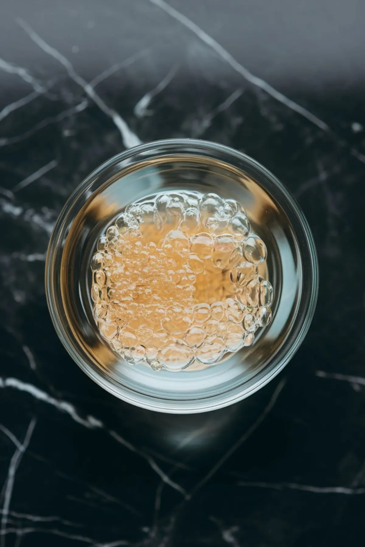 Close-up of bloomed gelatin in a glass bowl, showing translucent gelatin sheets or powder fully hydrated and expanded, sitting on a black marble surface with visible texture and detail.