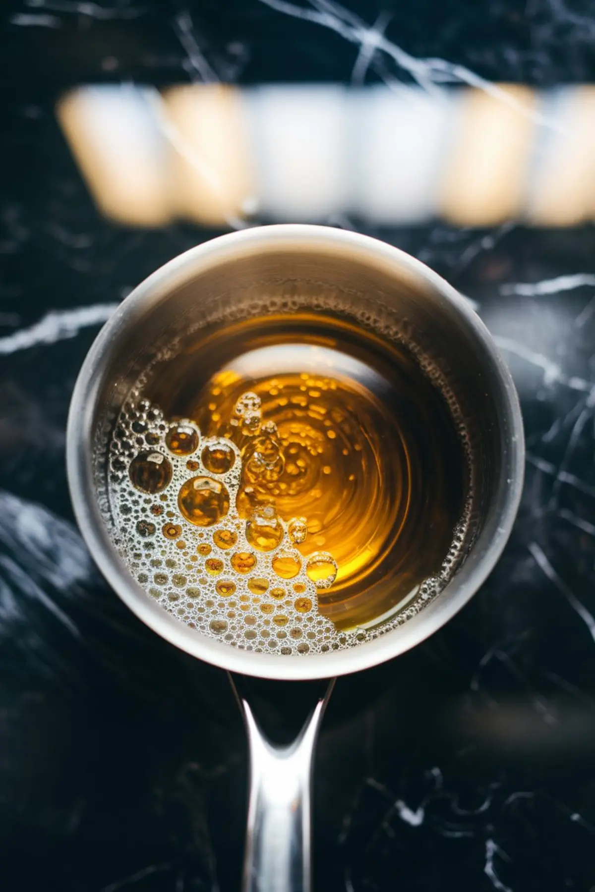 Overhead view of a saucepan with dissolved gelatin and sugar mixture, featuring golden liquid and bubbles on the surface, sitting on a glossy black marble countertop with reflective highlights.