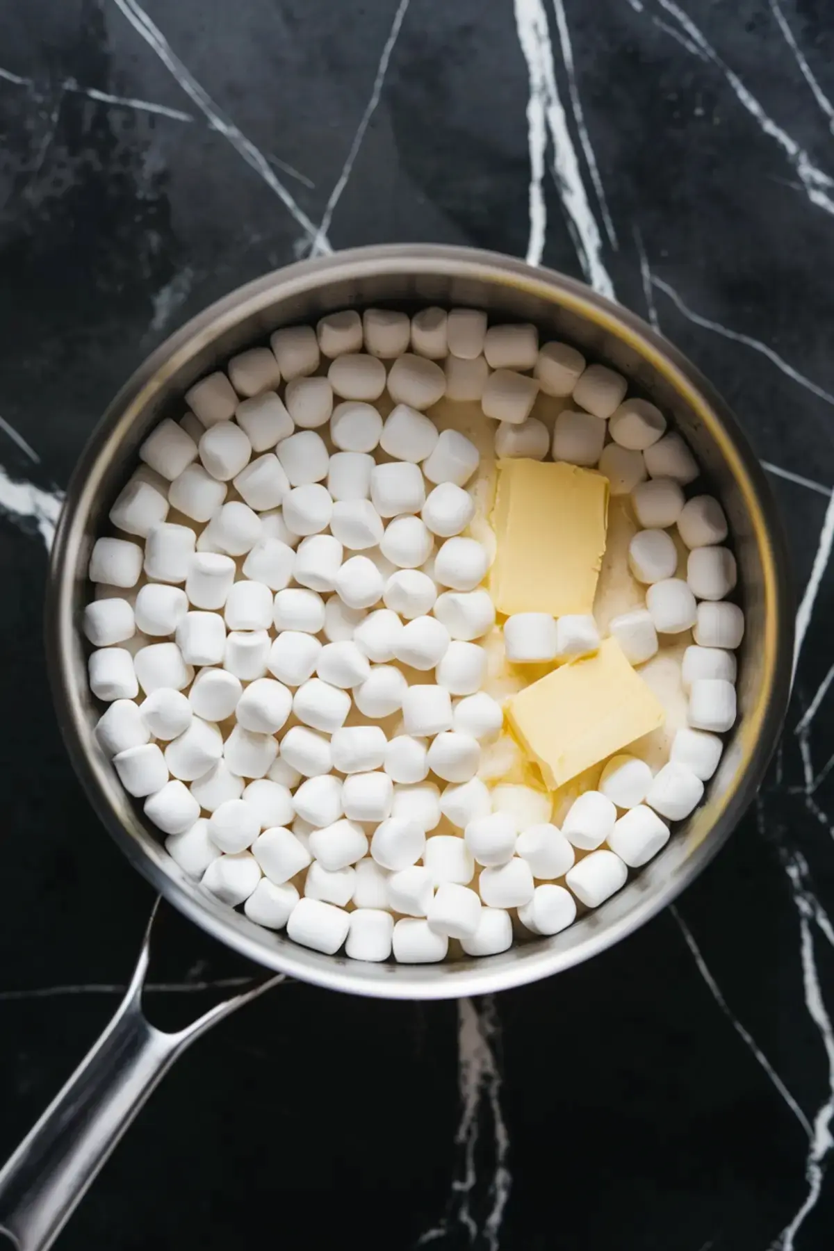 A saucepan filled with mini marshmallows and chunks of butter, set on a black marble countertop. The ingredients are beginning to melt together, creating the gooey base for Rice Krispie treats.