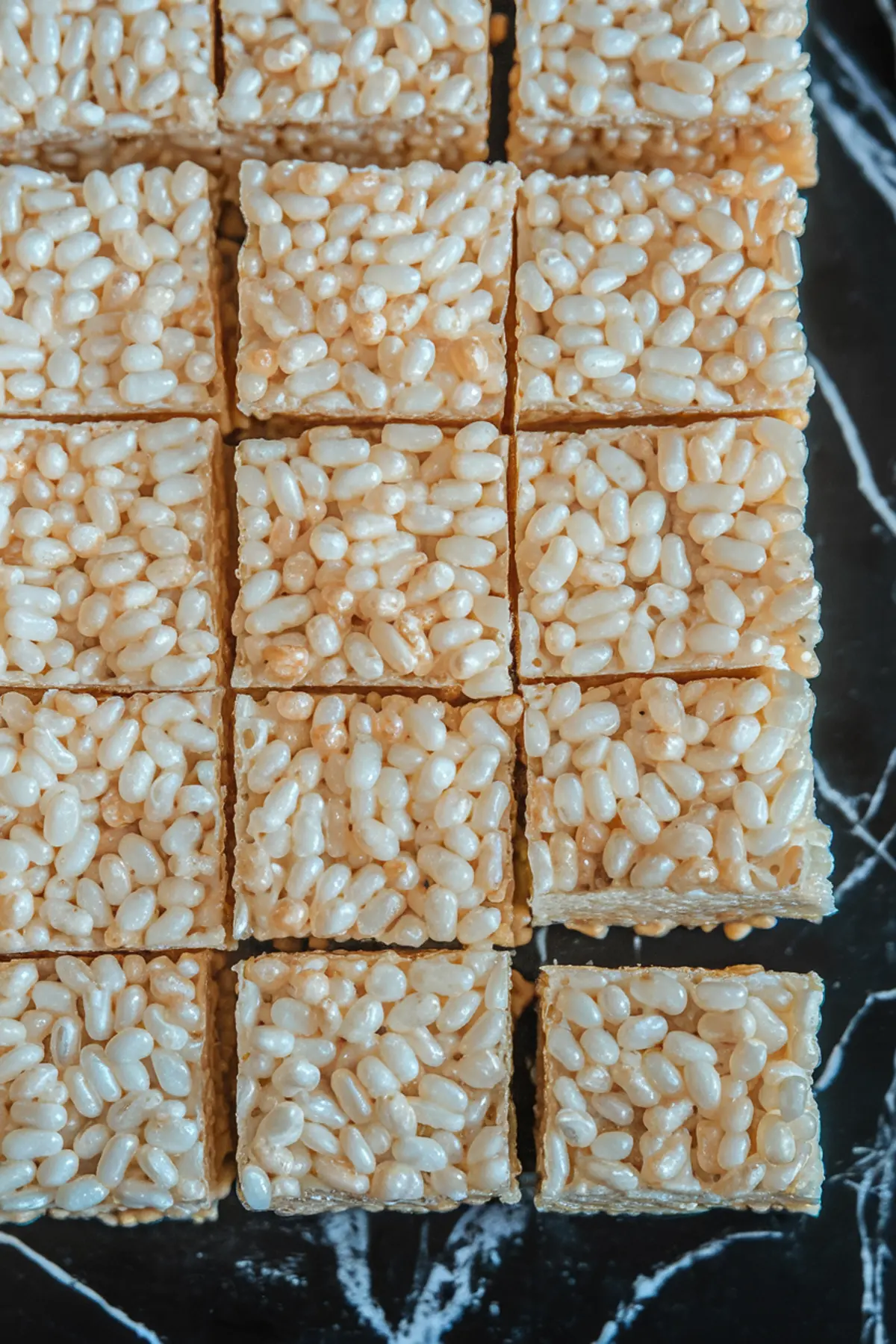 A batch of plain Rice Krispie treats cut into squares on a black marble surface. The puffed rice cereal is coated in a glossy marshmallow mixture, with clean edges and a uniform shape.