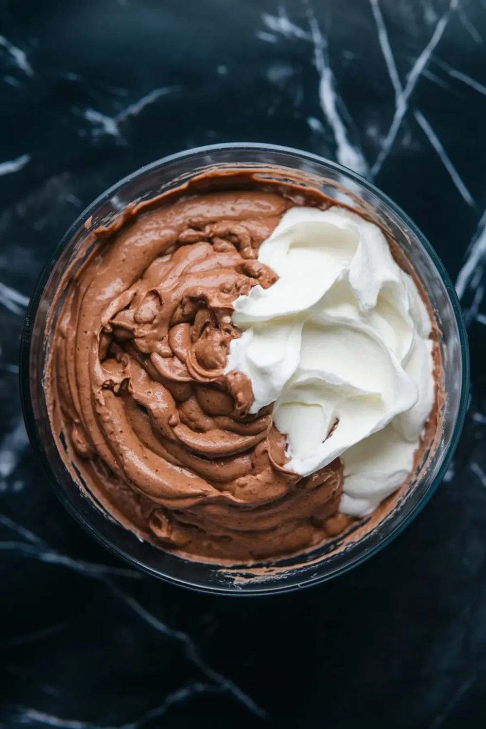 Glass bowl filled with smooth chocolate mousse and whipped cream, partially mixed, sitting on a black marble countertop; ideal visual for dessert preparation or no-bake recipe inspiration.