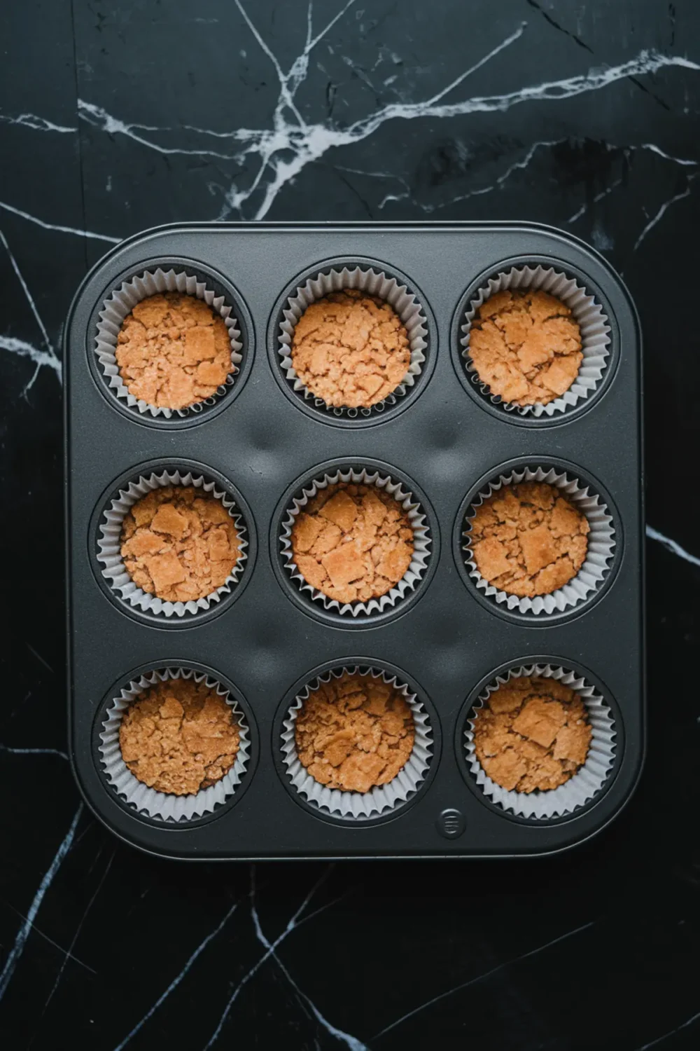 Non-stick muffin tin with nine graham cracker crust bases pressed into cupcake liners, ready for filling; set against a black marble background, showcasing a dessert prep stage.