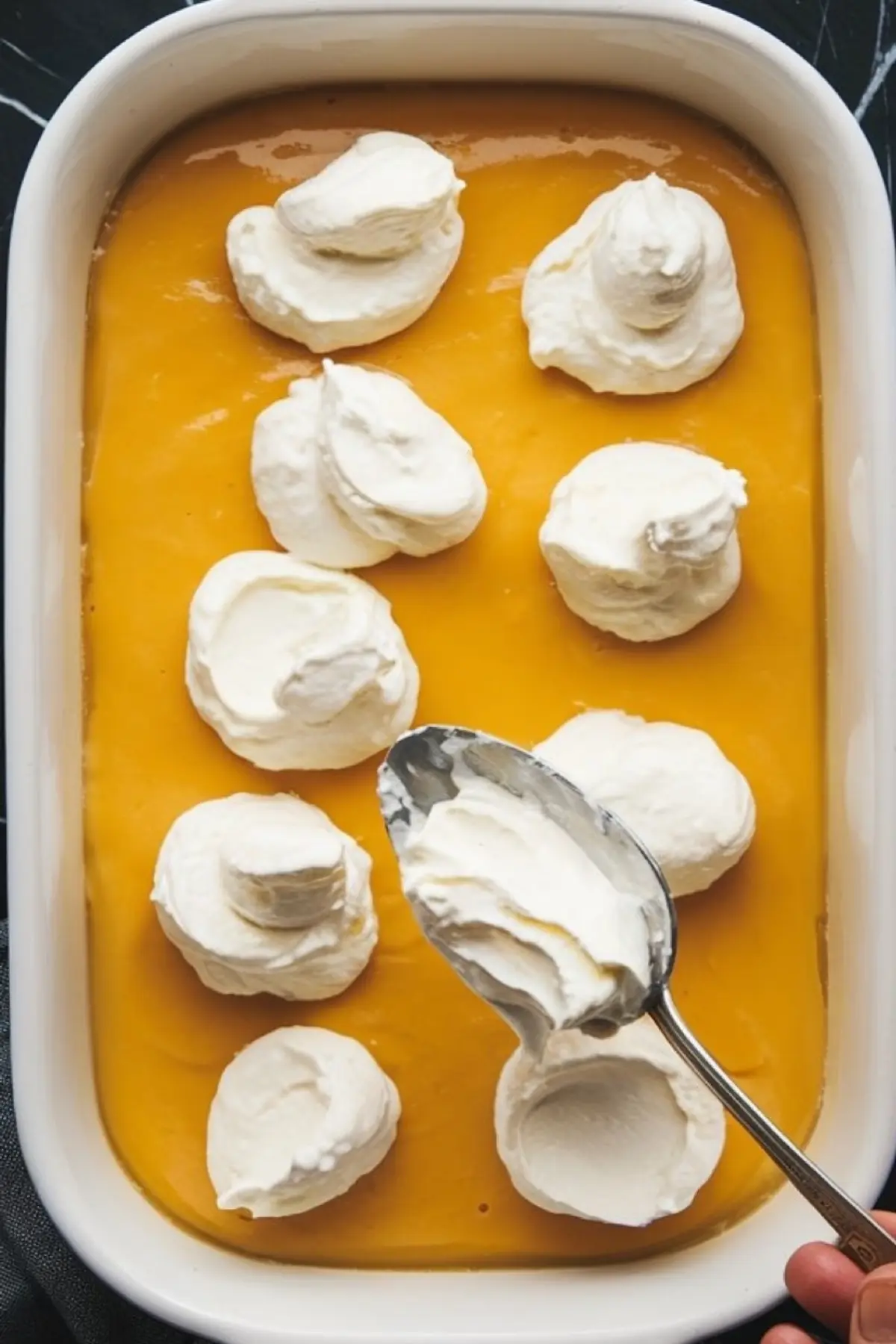 A spoon spreads dollops of whipped topping over a smooth lemon filling in a rectangular white baking dish, showing a step in assembling a lemon cream cheese dump cake.