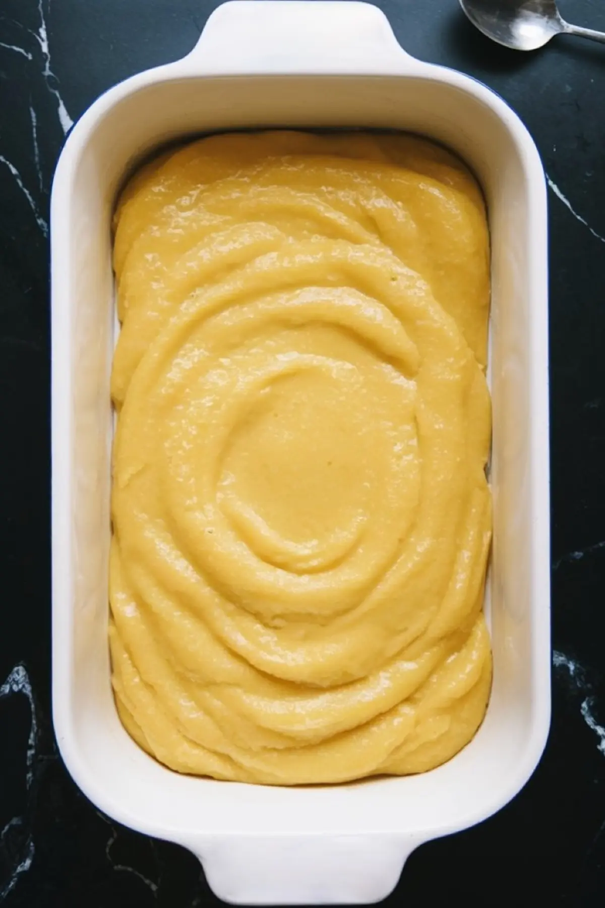Thick yellow cake batter is evenly spread in a white ceramic baking dish, forming the base layer for a lemon cream cheese dessert.