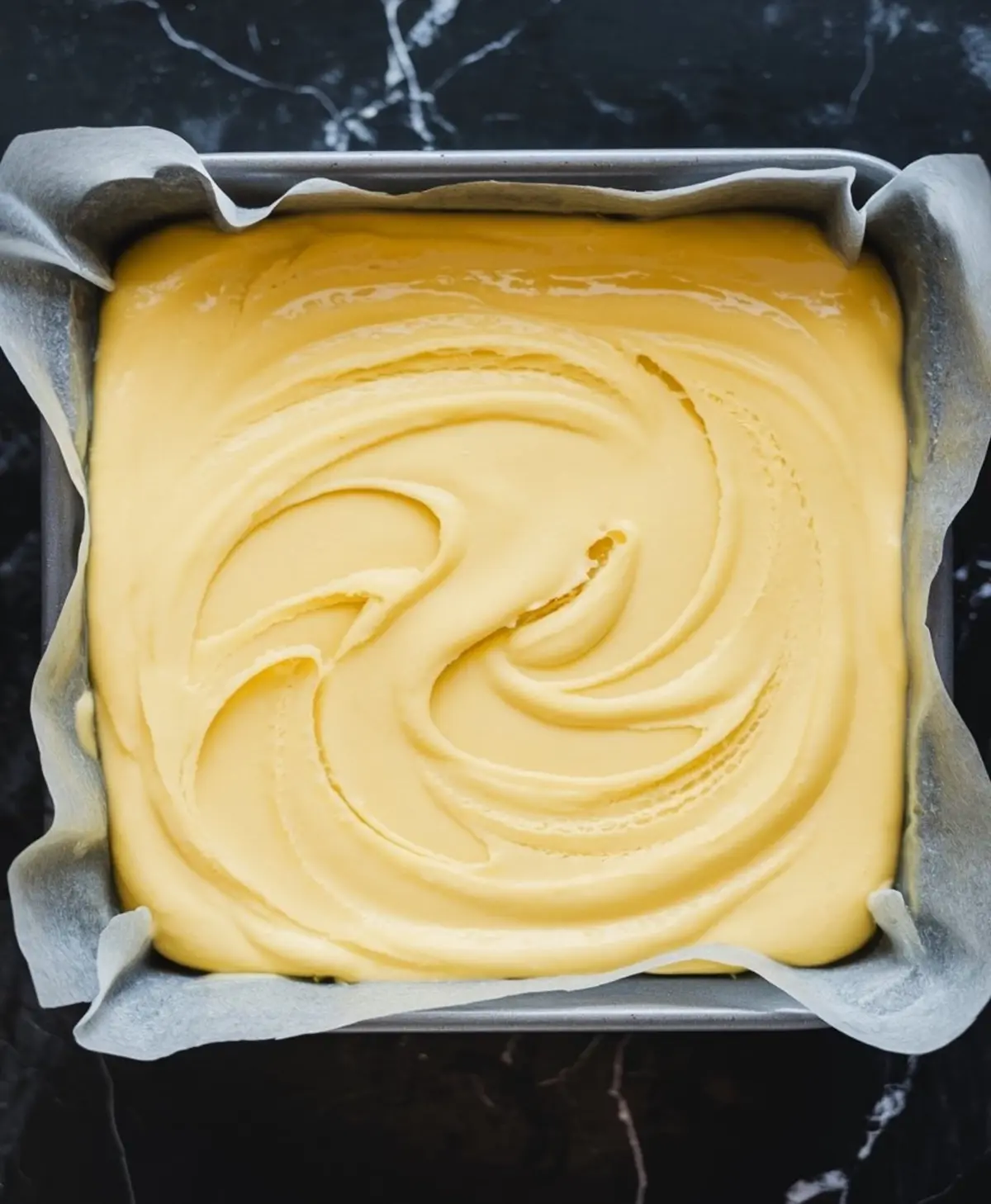 A square baking pan lined with parchment paper, filled with freshly poured lemon fudge mixture. The smooth surface features gentle swirls, giving the fudge a glossy and creamy appearance before setting.