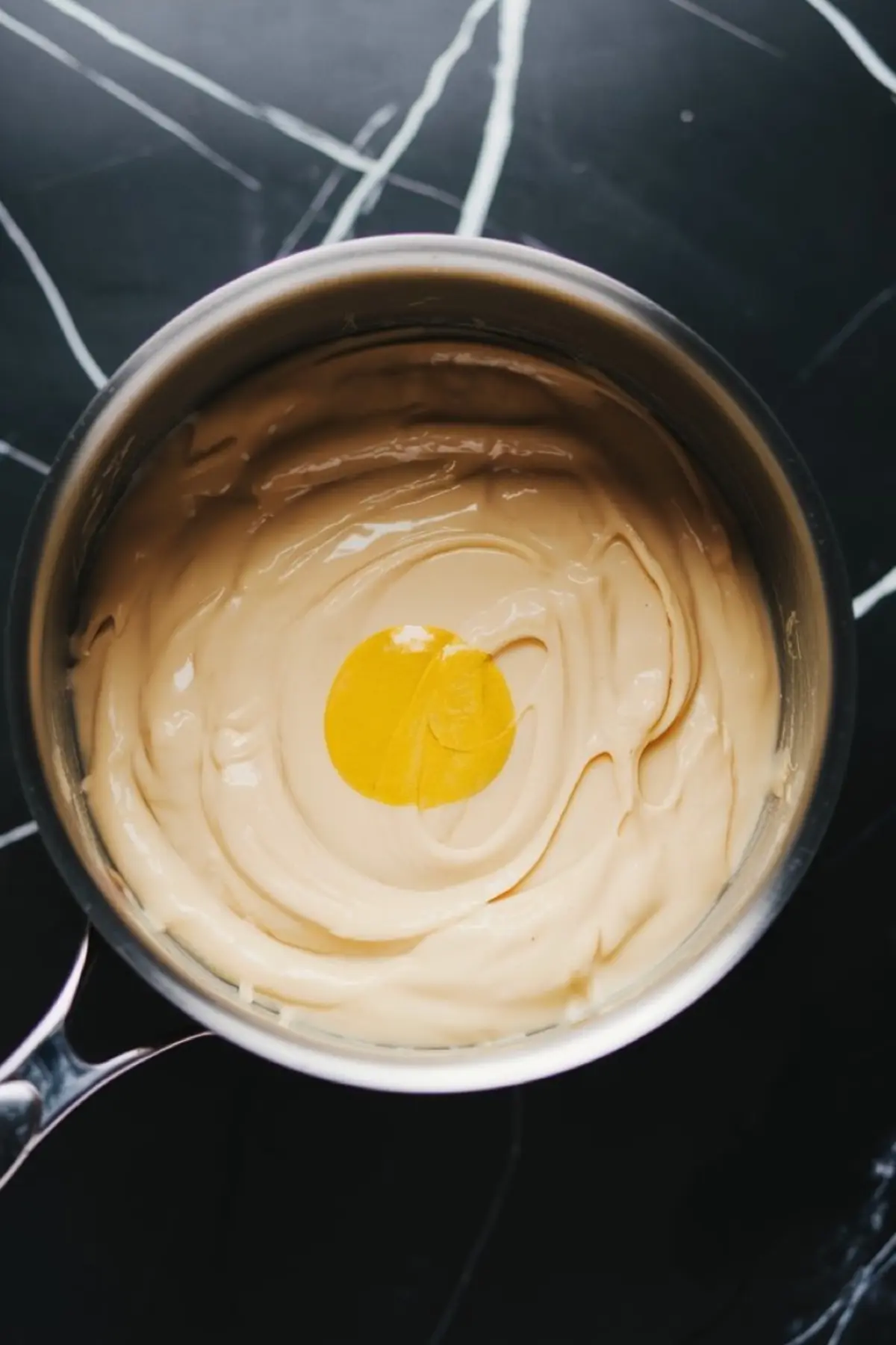 A saucepan with a creamy, melted white chocolate mixture, with a pool of bright yellow lemon extract in the center. The mixture is smooth and glossy, preparing to be combined for a rich lemon-flavored fudge.