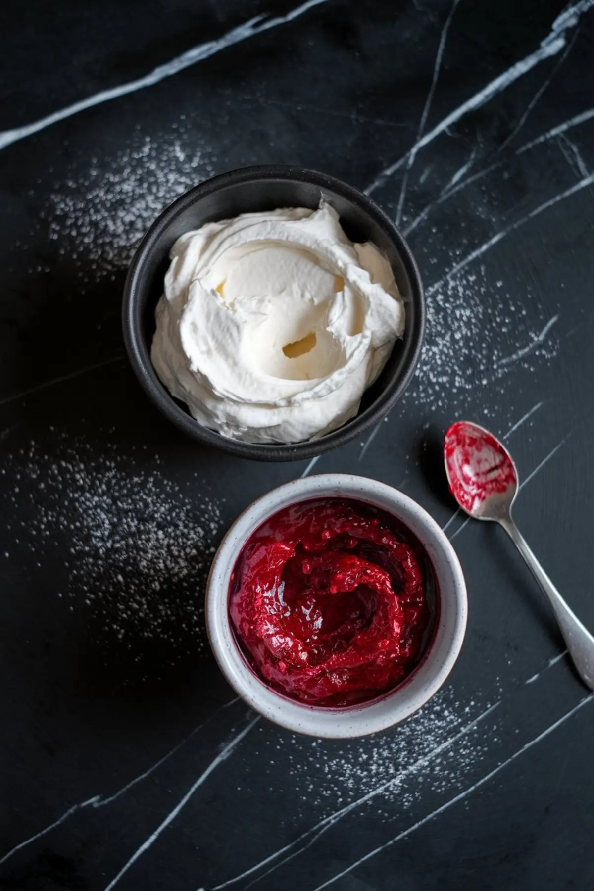 Overhead view of whipped cream cheese filling and homemade raspberry jam in black and white bowls on a black marble surface, with powdered sugar scattered and a jam-covered spoon beside the bowls.