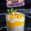 Glass of mango coconut chia pudding parfait topped with fresh mango chunks, shredded coconut, and a mint sprig, set against a black marble background.