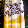 Mango lassi popsicle with bright orange and creamy white layers, sprinkled with pistachios and saffron, featuring bold “Mango Lassi Popsicles” text overlay.