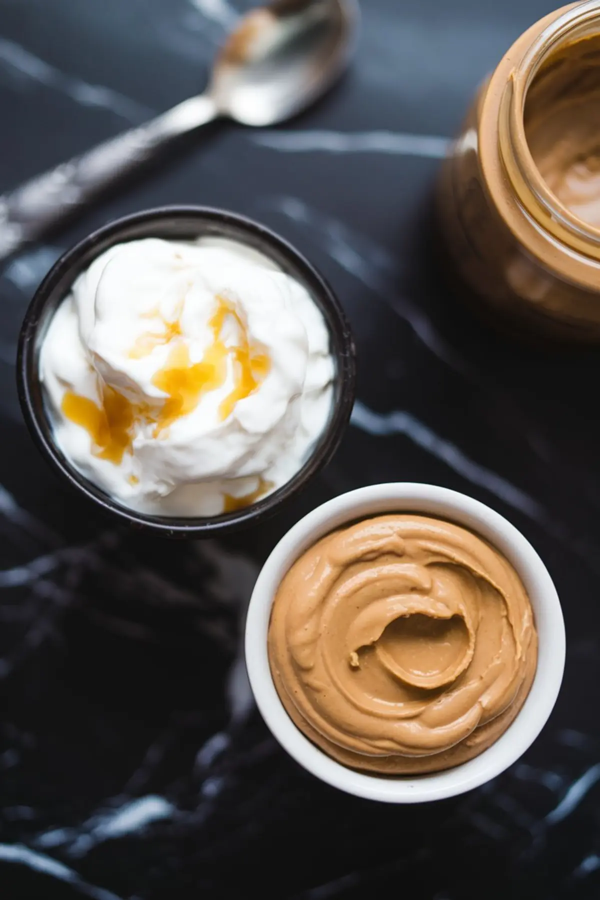 Overhead view of two small bowls on black marble, one with thick yogurt drizzled with honey, and one with creamy almond butter, alongside a spoon and glass jar in the background.
