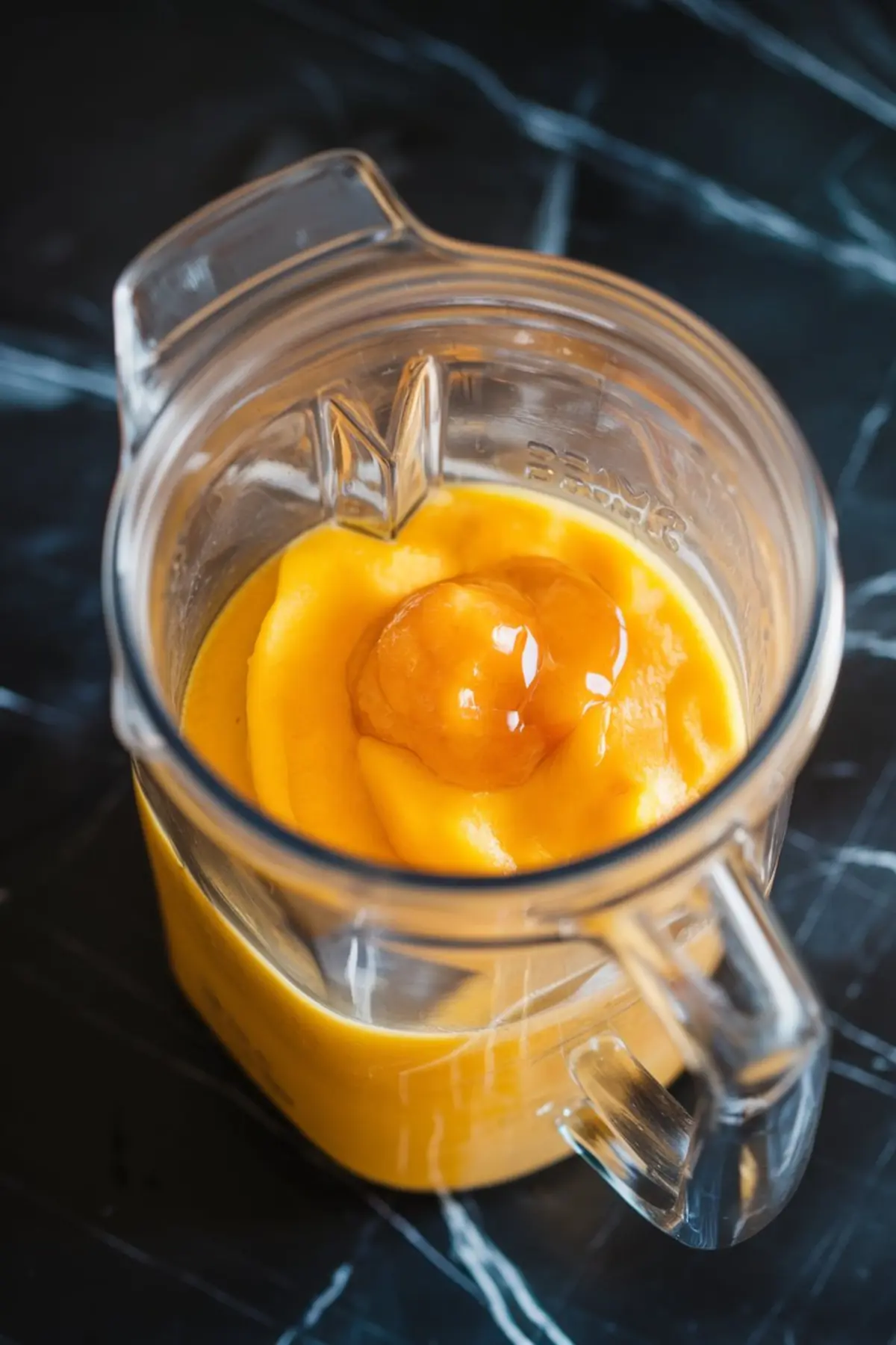 Clear blender filled with blended mango puree and honey on a dark marble surface, showcasing the thick and vibrant texture used for mango lassi popsicles.