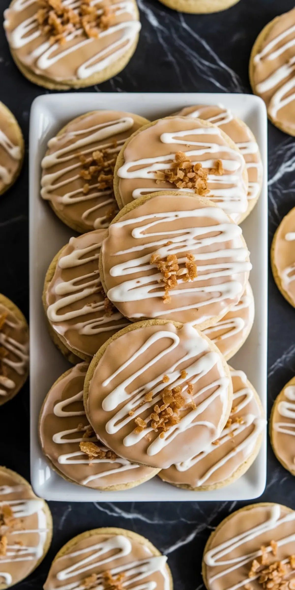 A top-down view of maple brown sugar cookies stacked on a white platter, surrounded by more cookies on a black marble surface. Each cookie is covered with a glossy maple glaze, white icing drizzle, and topped with caramelized brown sugar crumbles, showing a homemade rustic look.