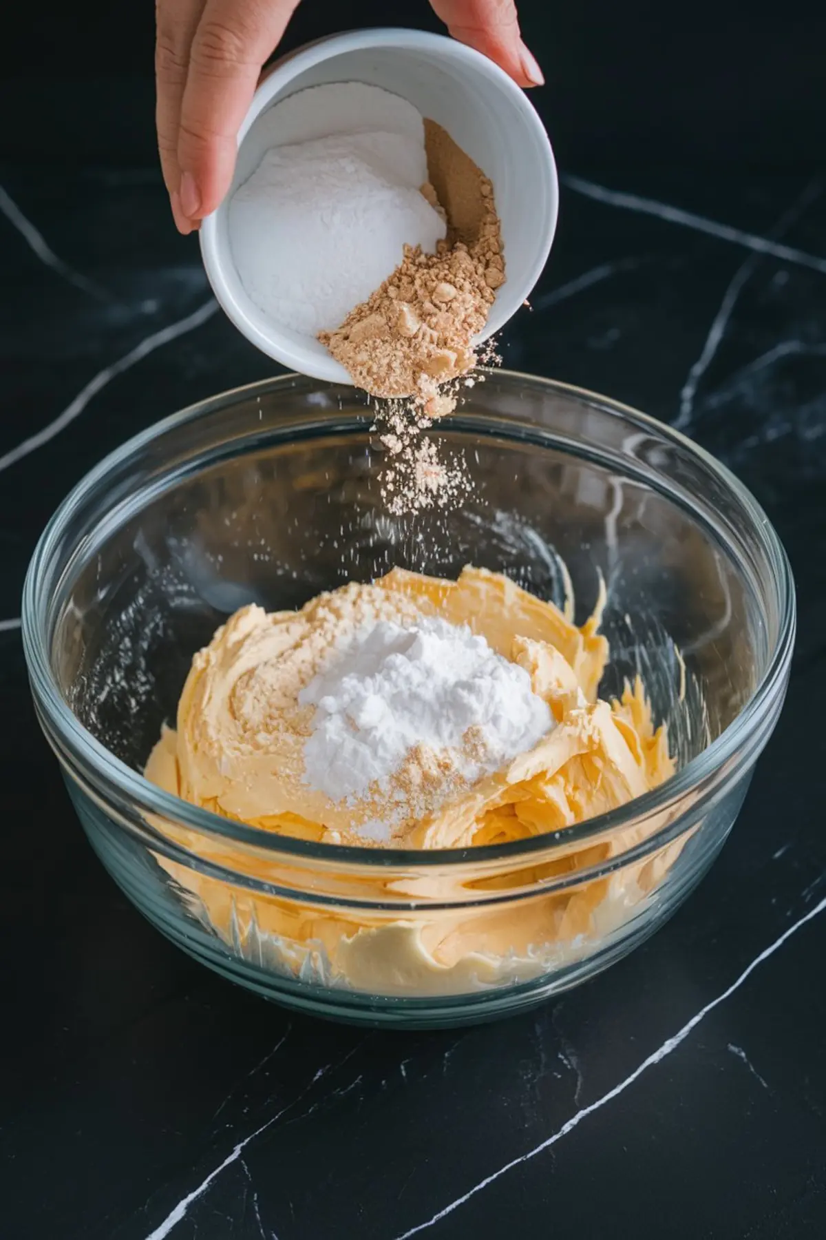 A close-up of a glass bowl filled with whipped butter, as powdered sugar and brown sugar are being poured in from a small white ramekin. Captured in motion, the image highlights the beginning of mixing cookie dough ingredients on a dark marble counter.