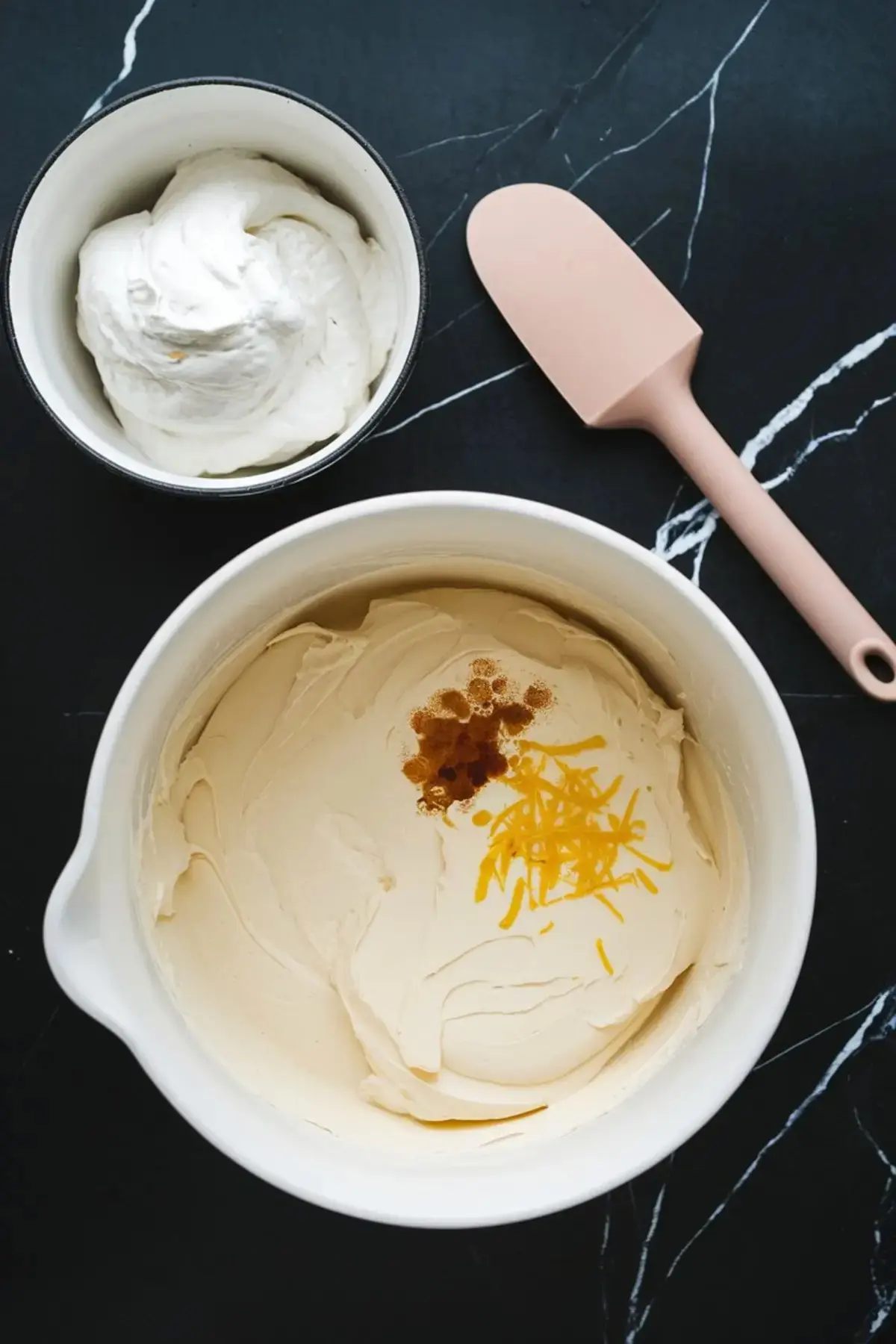 Mixing bowl filled with creamy cheesecake batter topped with vanilla extract and lemon zest, next to a bowl of whipped cream and a pink silicone spatula, on a black marble countertop.