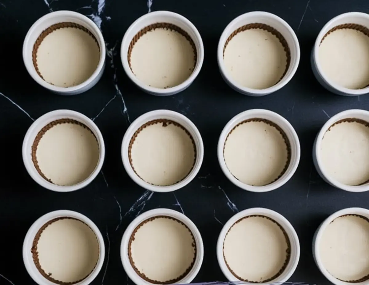 Image of twelve mini no-bake cheesecakes in white ramekins, showing smooth cheesecake filling and graham cracker crust, arranged in neat rows on a dark marble background.