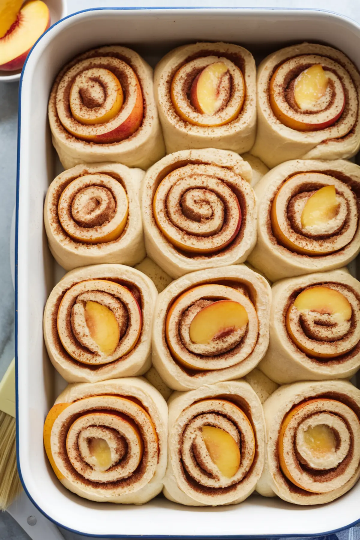 A white baking dish with unbaked peach cobbler cinnamon rolls, showing tightly rolled swirls of dough filled with cinnamon sugar and fresh peach slices nestled inside.