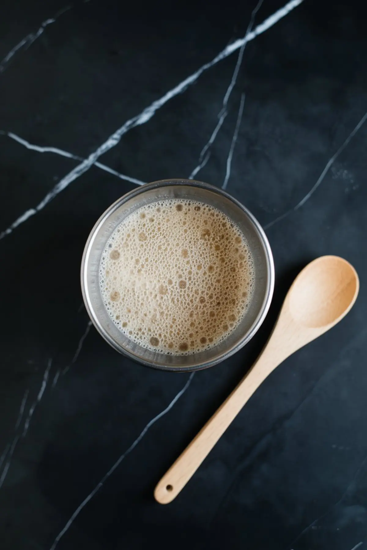 Activated yeast mixture in a metal bowl with a wooden spoon beside it, sitting on a dark marble countertop.