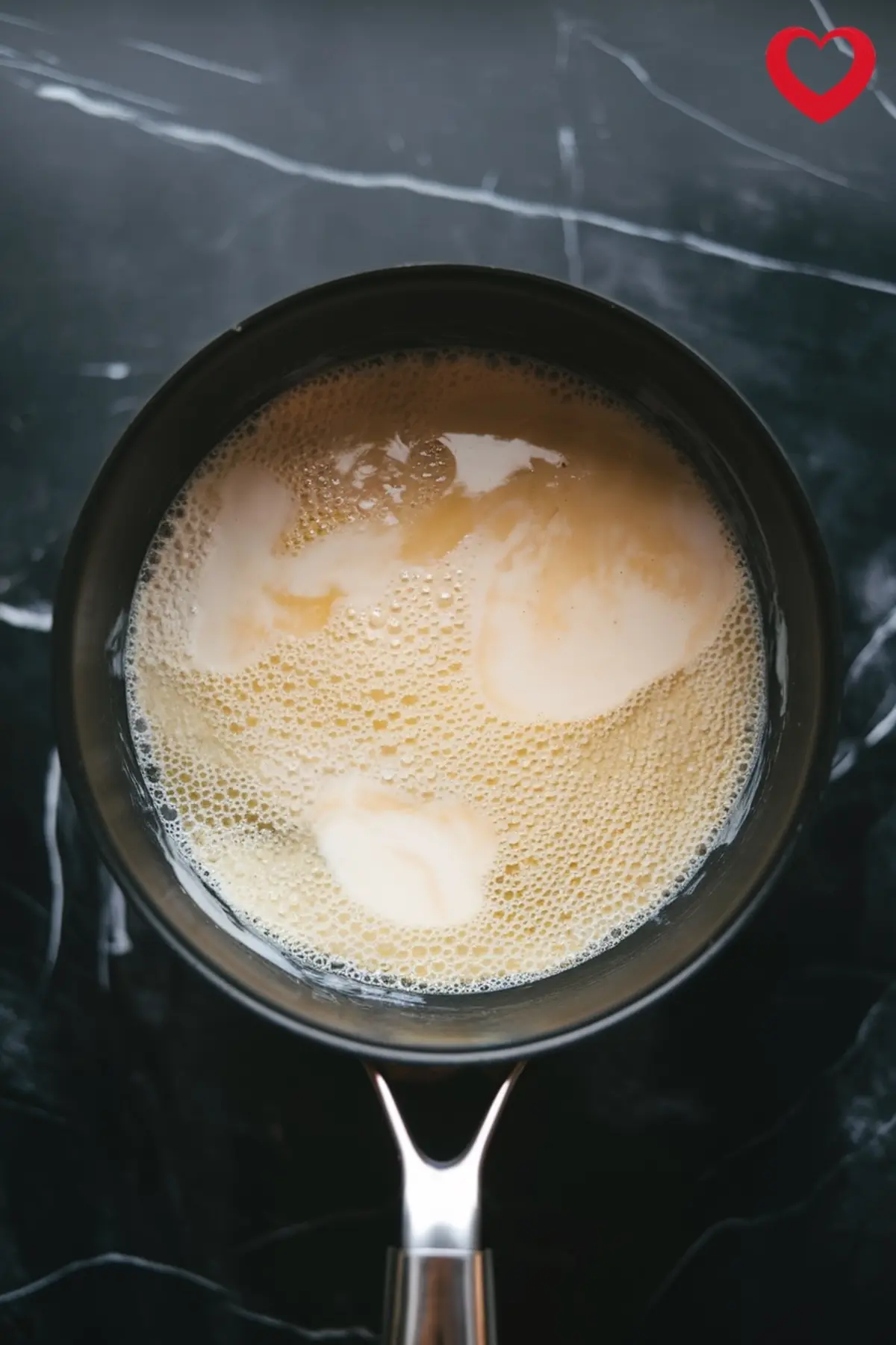 Top view of a saucepan filled with a foamy, creamy coconut mixture being heated on a black marble surface, part of the preparation for piña colada panna cotta.