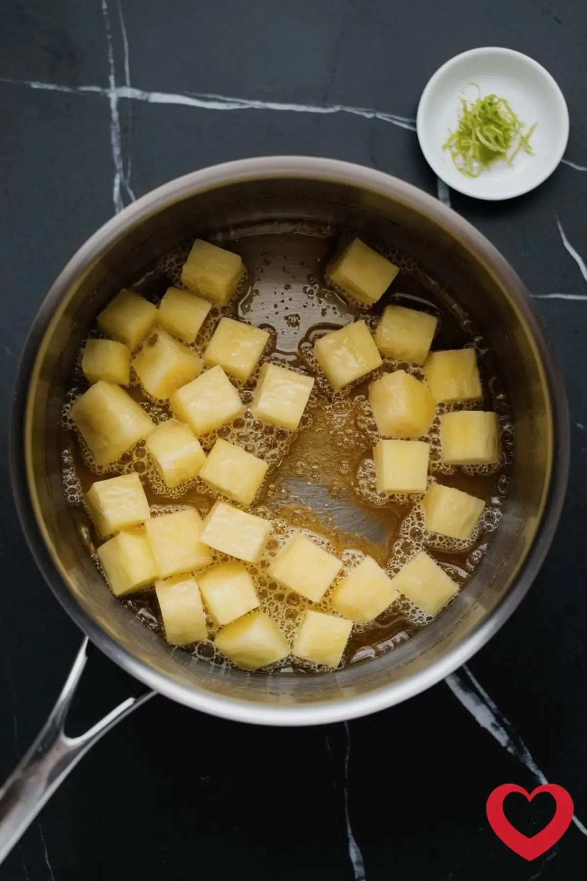 Cubed pineapple simmering in a saucepan with a bubbling golden syrup, with a small white dish of lime zest placed nearby on a black marble countertop.