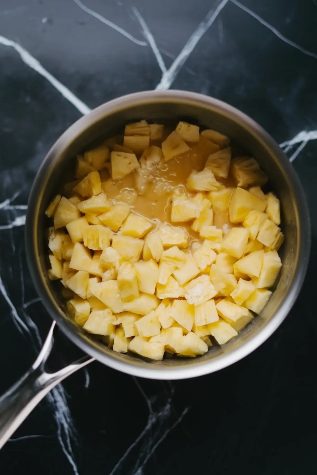 Saucepan filled with chopped fresh pineapple pieces in juice, sitting on a black marble countertop, ready to be cooked for the pineapple crumble bar filling.