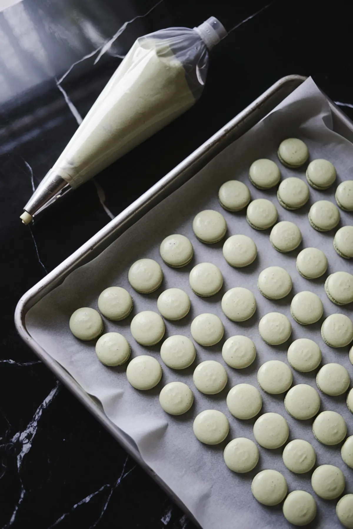 A baking tray lined with parchment paper holds neatly piped green macaron shells in uniform rows. A piping bag filled with macaron batter rests beside the tray, showcasing the delicate preparation process.