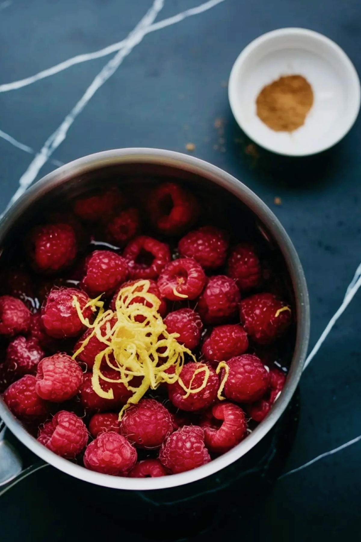 A saucepan filled with fresh raspberries and lemon zest on a dark countertop. A small dish of cinnamon is placed nearby, indicating the ingredients for the raspberry filling.