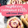 A close-up of raspberry cinnamon rolls with lemon glaze on a dark surface. The rolls are golden brown with vibrant raspberry filling swirled inside, drizzled with icing, and sprinkled with lemon zest. A decorative banner reads “Raspberry Cinnamon Rolls with Lemon Glaze.”