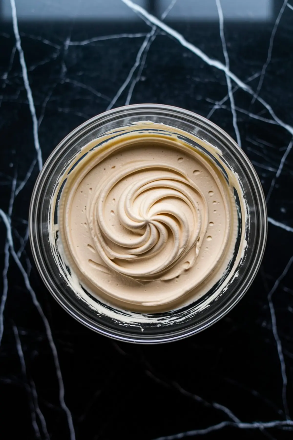  A glass bowl filled with creamy beige buttercream frosting, swirled into a smooth, decorative pattern, placed on a black marble surface.