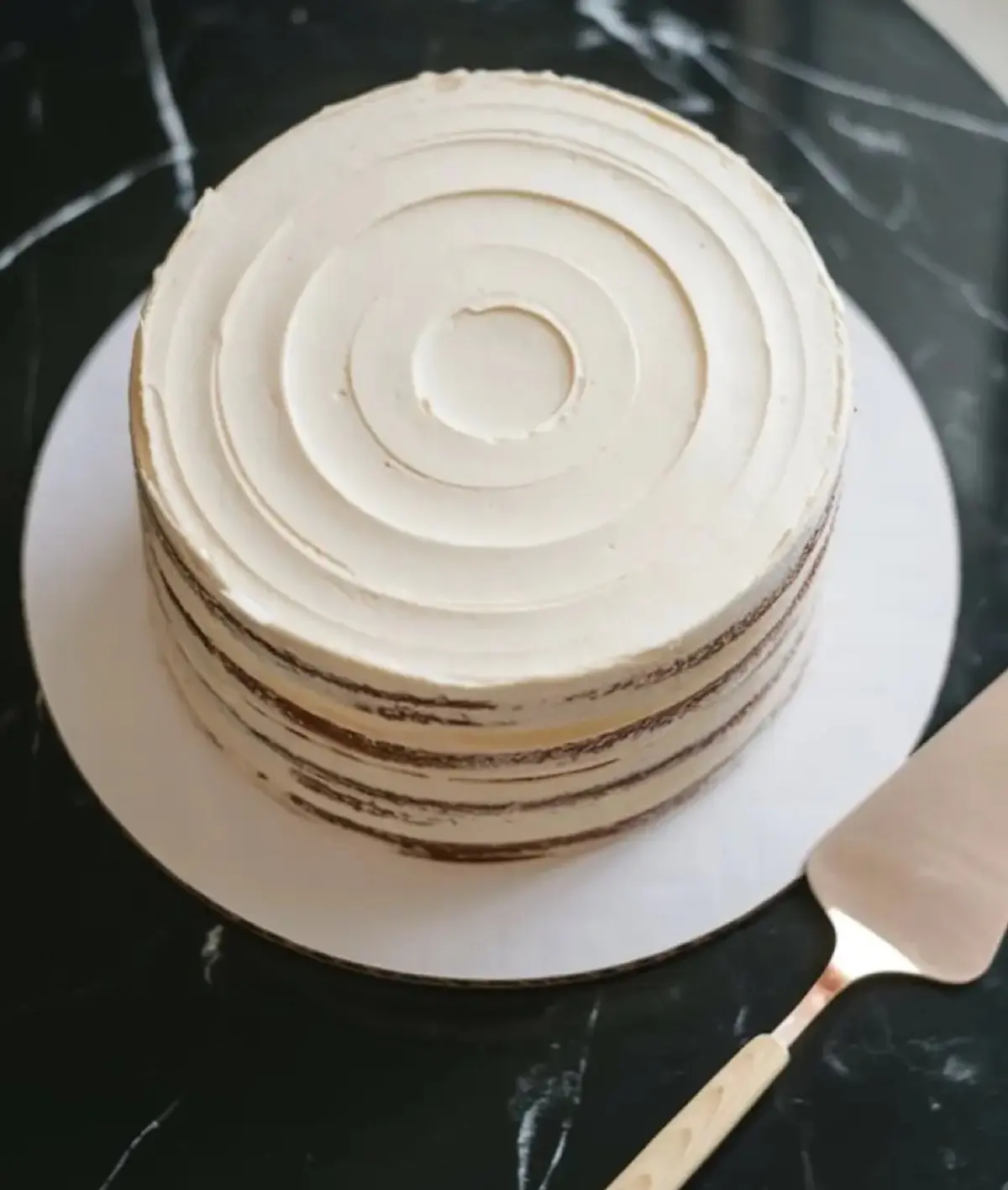 A minimalist layer cake with smooth white buttercream frosting, featuring concentric circular patterns on top and semi-naked edges revealing chocolate cake layers, placed on a white cake board with a cake server on a black marble surface.