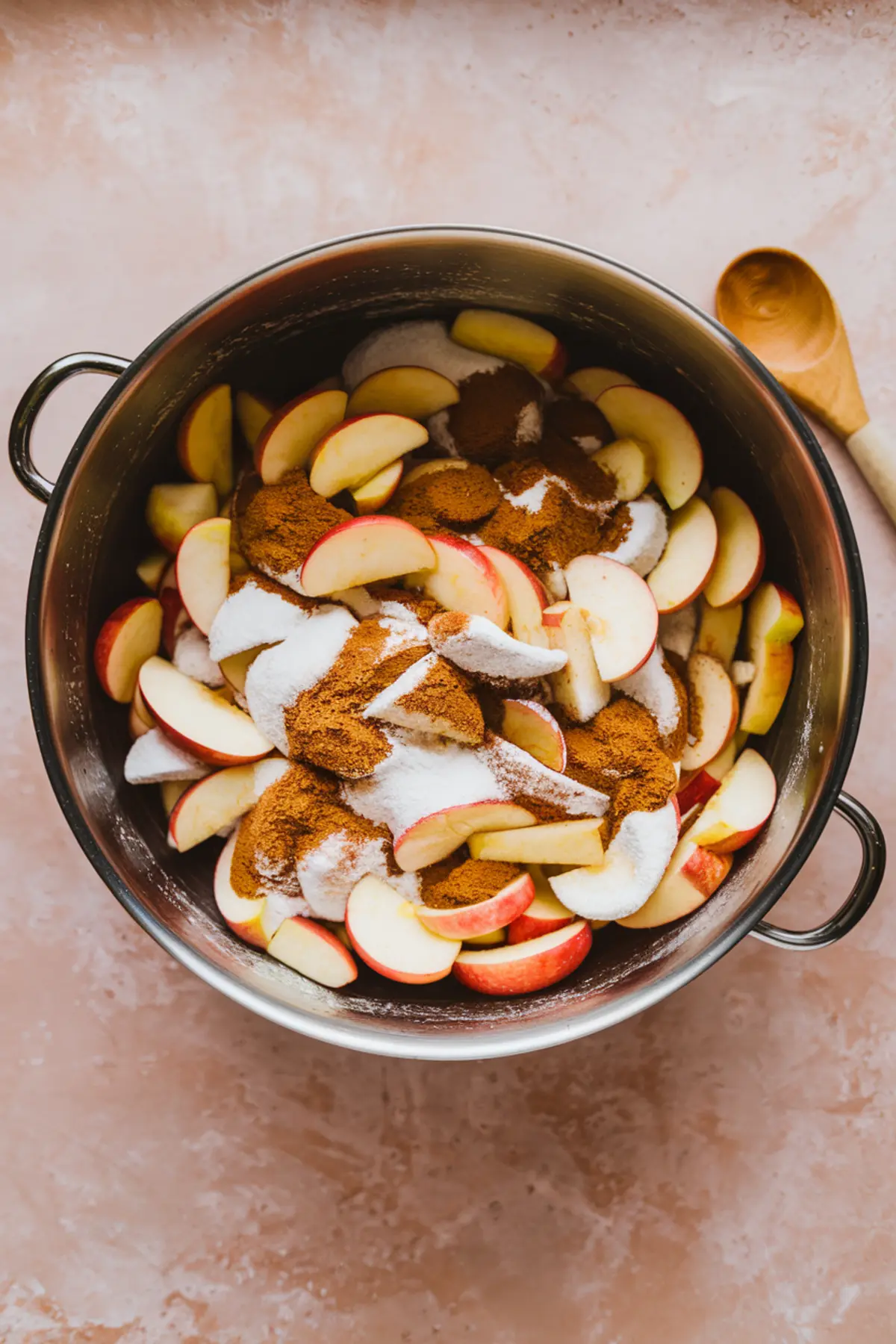 Large metal mixing bowl filled with fresh apple slices coated in granulated sugar, brown sugar, cinnamon, and spices, ready for baking into an apple pie.