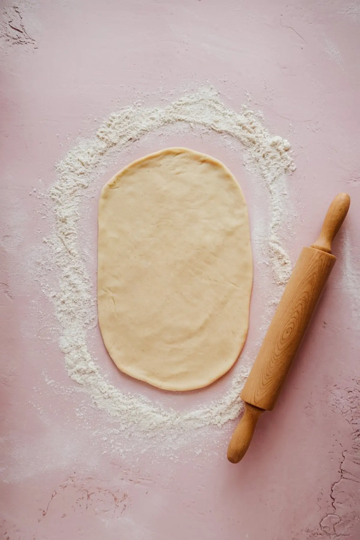 Oval sheet of pie crust dough rolled out on a pink floured surface, with a wooden rolling pin placed nearby.