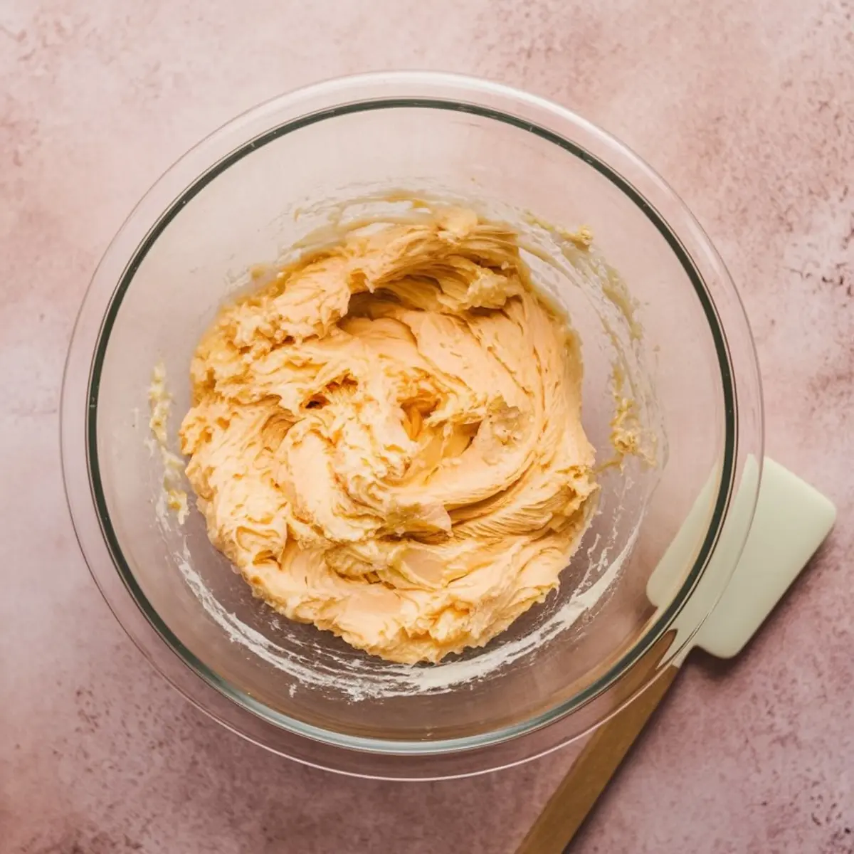 Overhead view of creamy sugar cookie dough in a glass mixing bowl with a rubber spatula beside it, placed on a pink countertop. Ideal for baking step-by-step visuals or cookie recipe process shots.