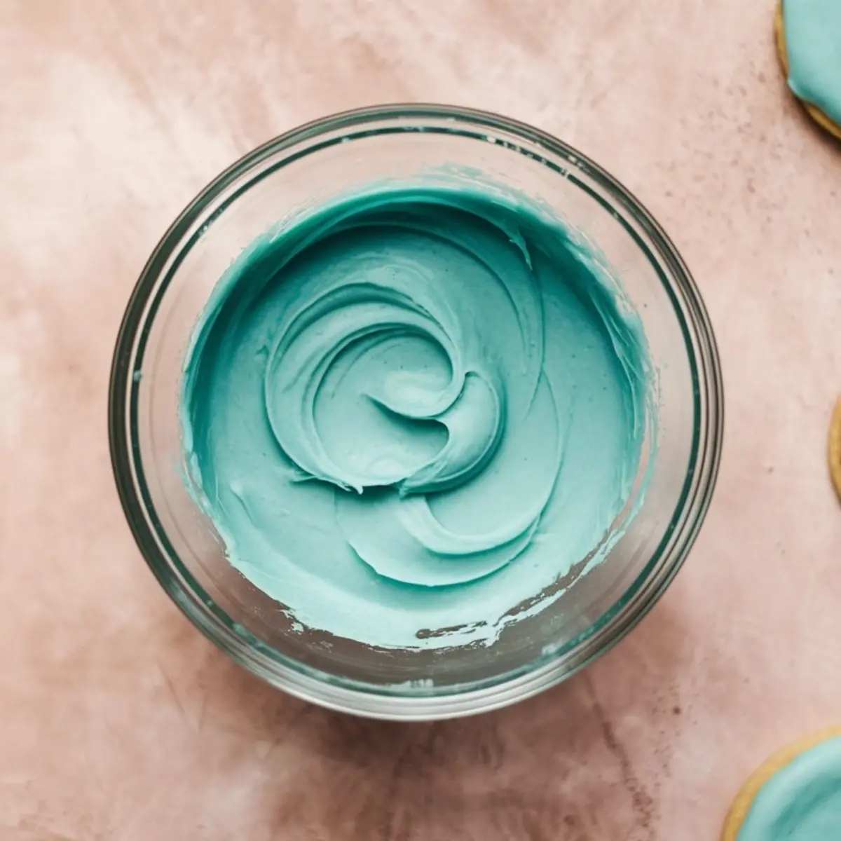 Smooth and glossy blue royal icing swirled in a glass bowl, placed on a pink countertop with cookie edges visible at the sides. Perfect for cookie decorating tutorials or icing consistency reference.