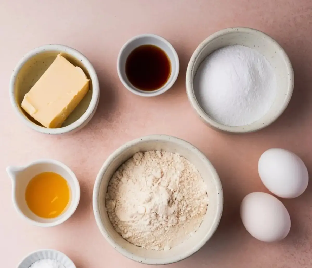 Flat lay of baking ingredients including flour, sugar, eggs, butter, vanilla extract, oil, baking powder, and water arranged on a pink surface. Great for recipe ingredient shots or baking prep imagery.