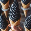 Hand holding a black charcoal soft serve ice cream cone, surrounded by four cones placed in a circular tray. The rich swirl of the soft serve contrasts with the golden waffle cone. Captures trendy dessert styling with bold color and texture.