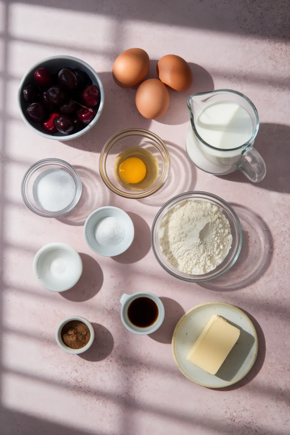 Overhead view of cherry clafoutis ingredients on a pink surface, including fresh cherries, eggs, milk, flour, sugar, butter, cinnamon, vanilla extract, and baking powder in various bowls and containers.
