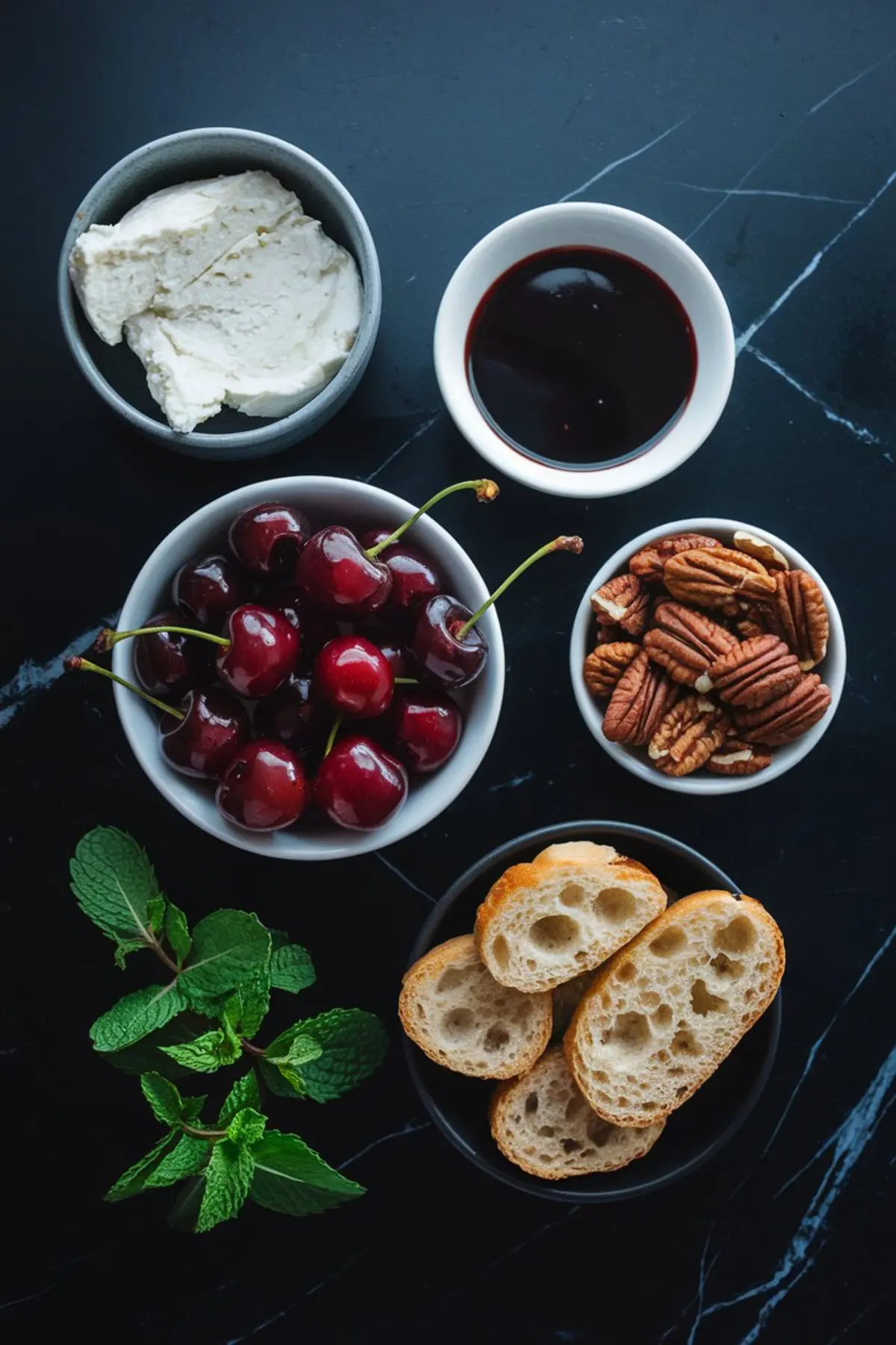 Top view of ingredients for cherry balsamic goat cheese dip on black marble, including fresh cherries, creamy goat cheese, pecan halves, balsamic glaze, mint leaves, and sliced baguette bread.