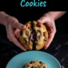 Close-up of hands splitting a gooey chocolate chip cookie over a teal plate with another whole cookie resting below.