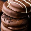 A close-up stack of chocolate-covered sandwich cookies featuring different toppings, including white chocolate drizzle and crushed cookies, set against a black background.